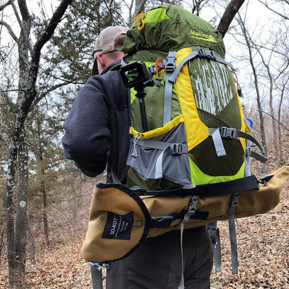 Man in the woods with backpack on his back.  Tan GO-KOT camping cot attached to the bottom of the backpack.