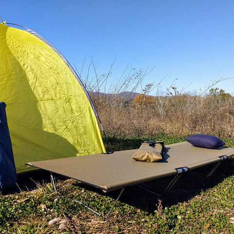 GO-KOT Tan Camping Cot and Snugpak Travel Pillow near yellow tent in a field.
